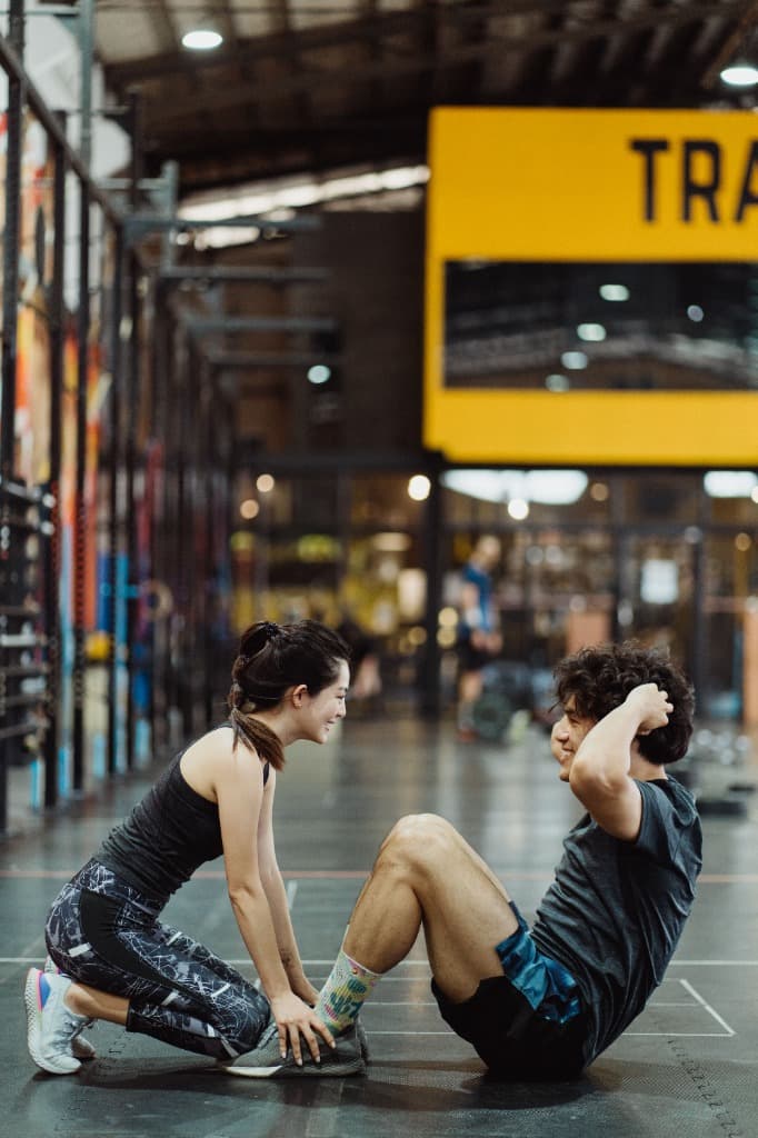 Two gym partners laughing, one holding the other's feet during sit-ups